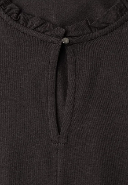 Schwarzes Shirt, Rundhalsausschnitt mit Rüsche, kleiner Schlitz mit Knopf und Bindeband.
