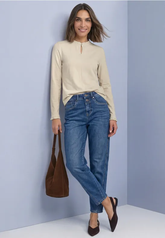 Frau in beiger Bluse, blauen Jeans, brauner Tasche und dunklen Loafers vor hellgrauer Wand, lächelnd.