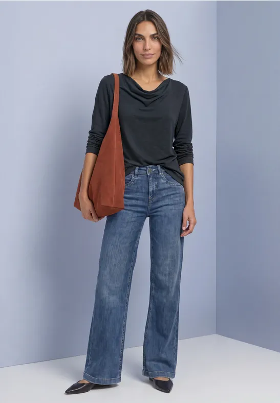 Vrouw tegen blauwe achtergrond met zwarte top, wijdvallende jeans, zwarte schoenen en een grote bruine tas.