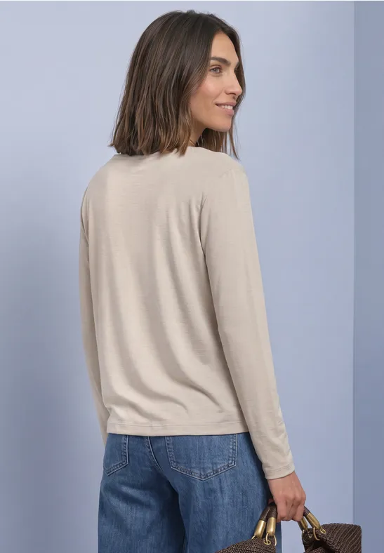 Vrouw in beige longsleeve en spijkerbroek, zijwaarts staand, met handtas voor lichte achtergrond.