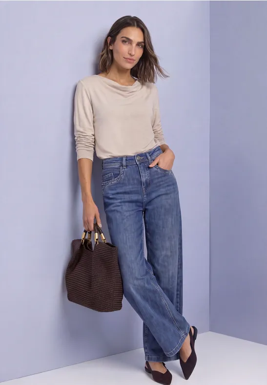 Vrouw in lichte trui en wijd jeans met handtas in de hand, ontspannen poserend voor een paarse muur.