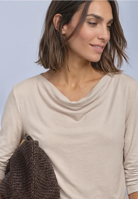 Vrouw met halflang bruin haar in een beige shirt met watervalhals draagt een gevlochten tas.