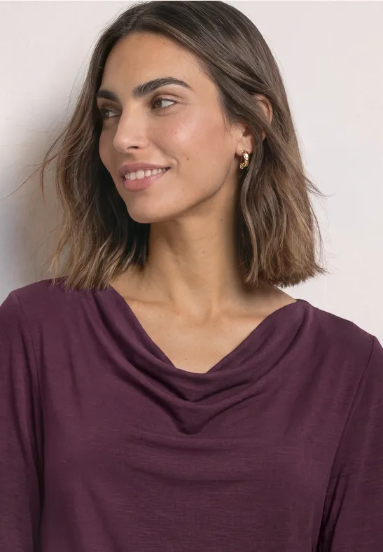 Een glimlachende vrouw met halflang bruin haar draagt een violet, los vallend shirt en kijkt iets opzij.