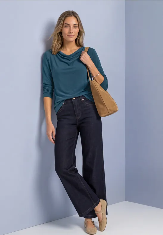Vrouw in groenblauw 3/4-mouw shirt, wijde donkere jeans, beige slippers en schoudertas tegen lichte achtergrond.