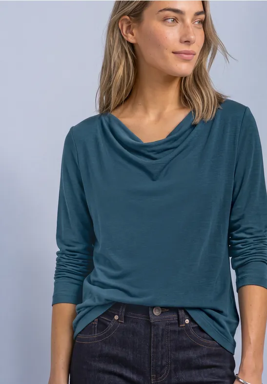 Vrouw in blauwgroene top met watervalhals en donkere spijkerbroek kijkt opzij
