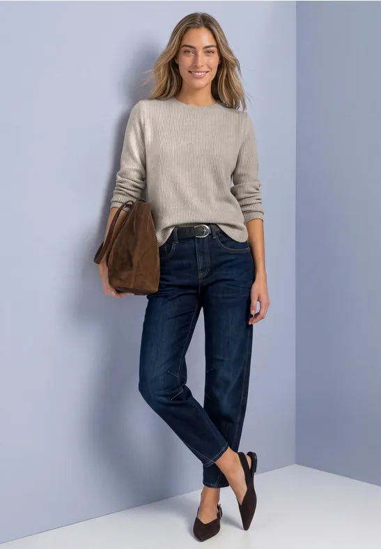 Une femme porte un pull, un jean foncé, des ballerines noires et tient un sac.