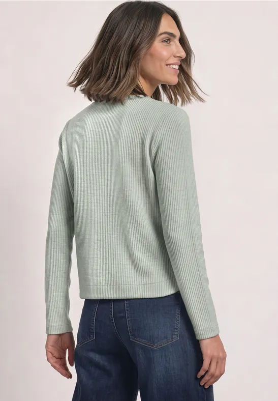 Femme de dos en pull vert clair et jean bleu, elle tourne la tête en souriant légèrement.