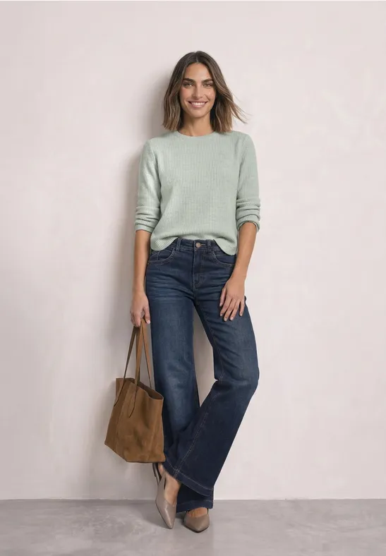 Une femme en pull vert, jean large, talons et sac en suède sur fond neutre.