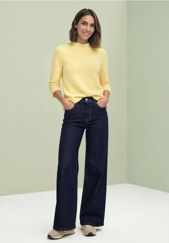 Eine Frau in gelbem Pullover und weiten dunklen Jeans steht vor hellem Hintergrund.