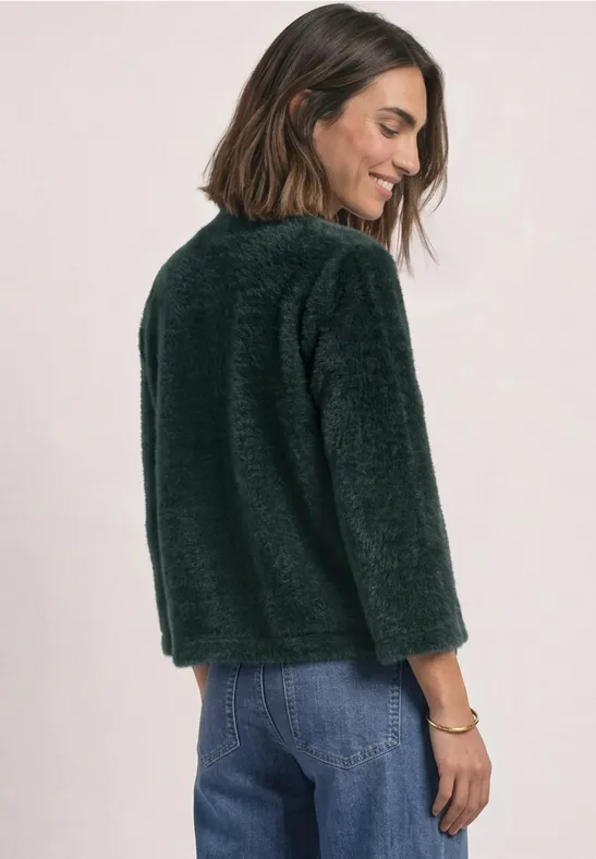 Femme aux cheveux mi-longs bruns porte un pull vert en peluche et un jean bleu, sourit de profil.