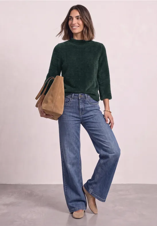 Femme en chemisier vert et jean large, tenant un sac brun, devant un fond clair.