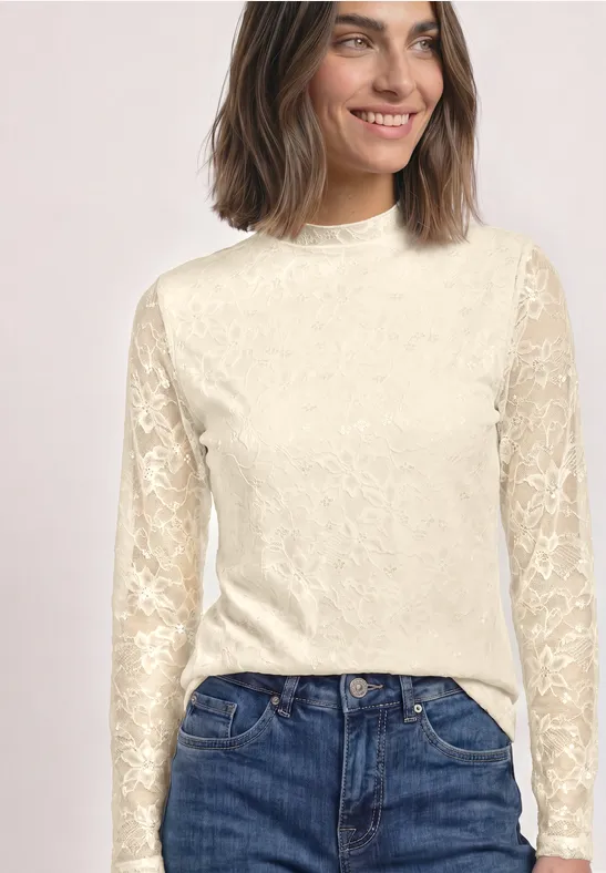 Vrouw draagt crèmekleurige kanten blouse met lange mouwen en een blauwe spijkerbroek.