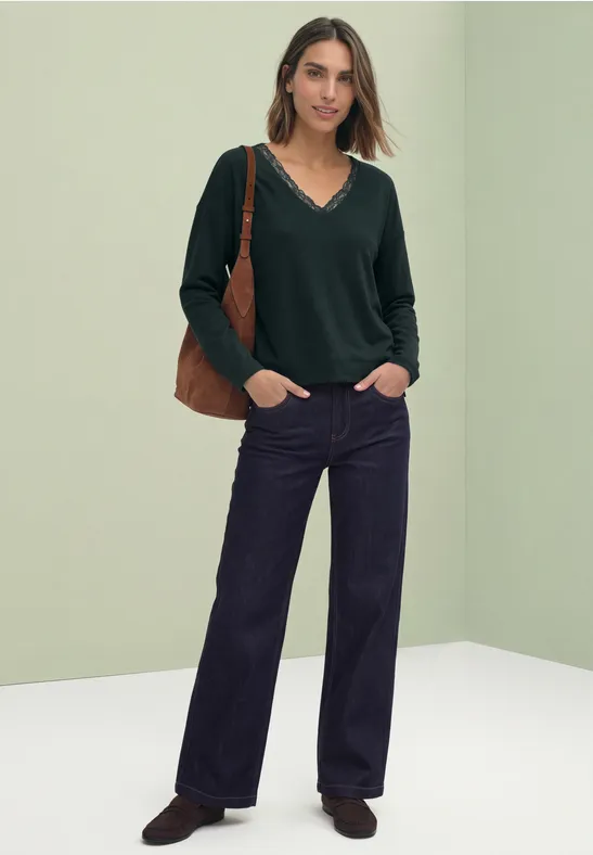 Vrouw in groene trui, wijde jeans en bruine tas, staand voor een lichtgroene achtergrond.