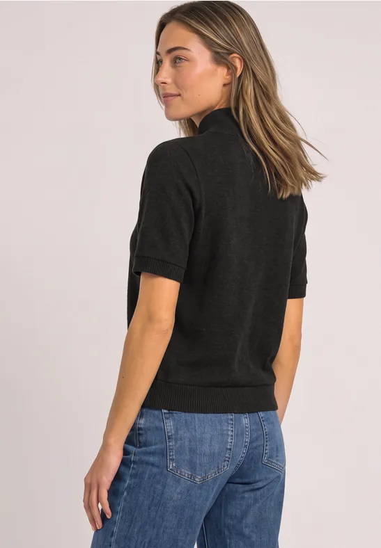 Frau in schwarzem Kurzarm-Top und blauen Jeans steht seitlich, Blick über die Schulter.
