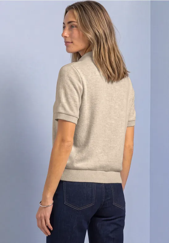 Frau in beigeem Kurzarm-Pullover und blauen Jeans im seitlichen Profil vor hellem Hintergrund.