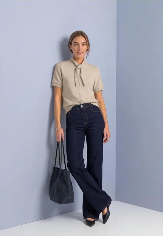 Frau in beiger Bluse mit Schluppe, dunkelblaue Schlagjeans und schwarzer Tasche vor pastellfarbener Wand.