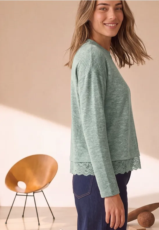 Eine Frau im Profil trägt ein grünes Langarmshirt mit Spitzenbesatz am Saum und dunkle Hosen.