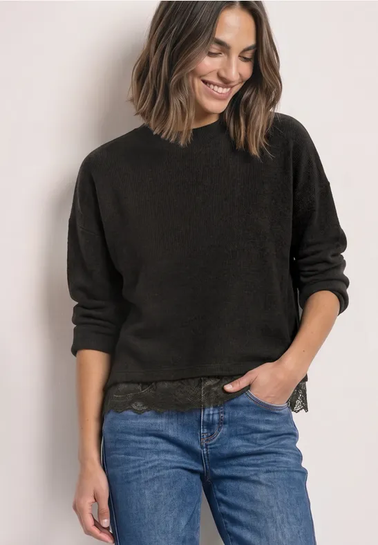 Frau mit schwarzem Pullover und Jeans steht lächelnd, eine Hand in der Hosentasche.
