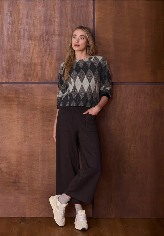 Femme en pull jacquard, pantalon large et baskets, posant devant une cloison en bois, main en poche.