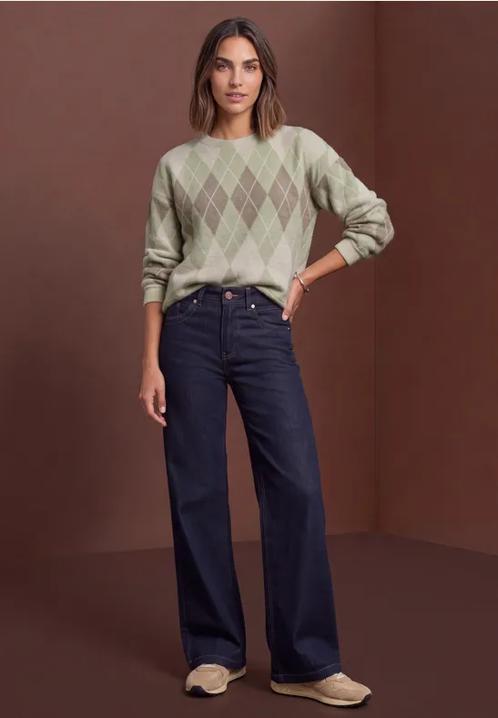 Frau in grünem Rautenmuster-Pullover, weiten Jeans und hellen Slippers vor braunem Hintergrund.