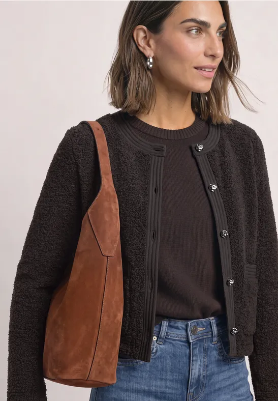 Frau trägt dunkelbraunes Strickjäckchen, schwarze Bluse, blaue Jeans und braune Wildleder-Tasche.
