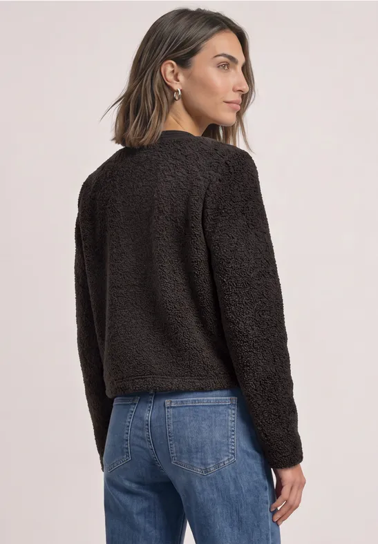 Frau mit kurzen Haaren trägt schwarze Teddy-Fleecejacke und blaue Jeans, schaut seitlich nach rechts.