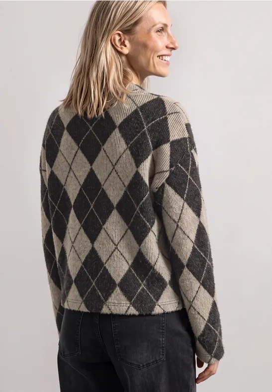 Een glimlachende blonde vrouw van achteren, in een beige-zwarte ruitjestrui en donkere jeans.