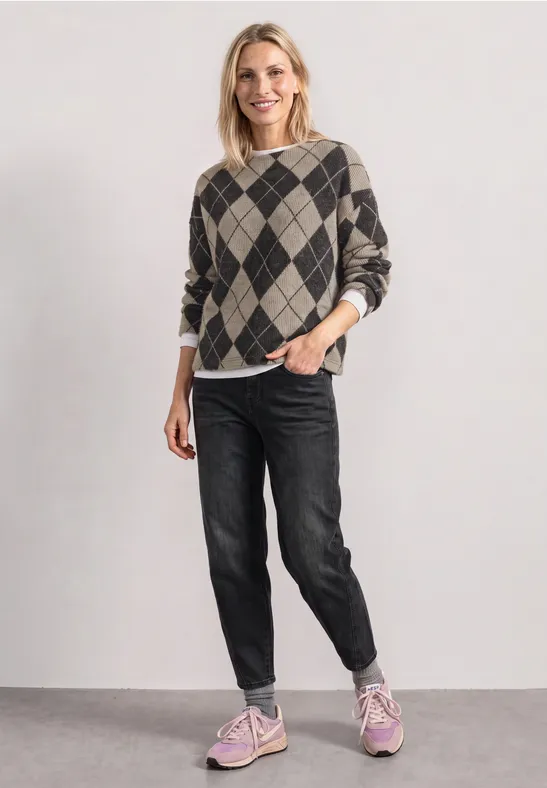 Vrouw lacht in een geruite trui, zwarte jeans en roze sneakers, staand voor een neutrale achtergrond.