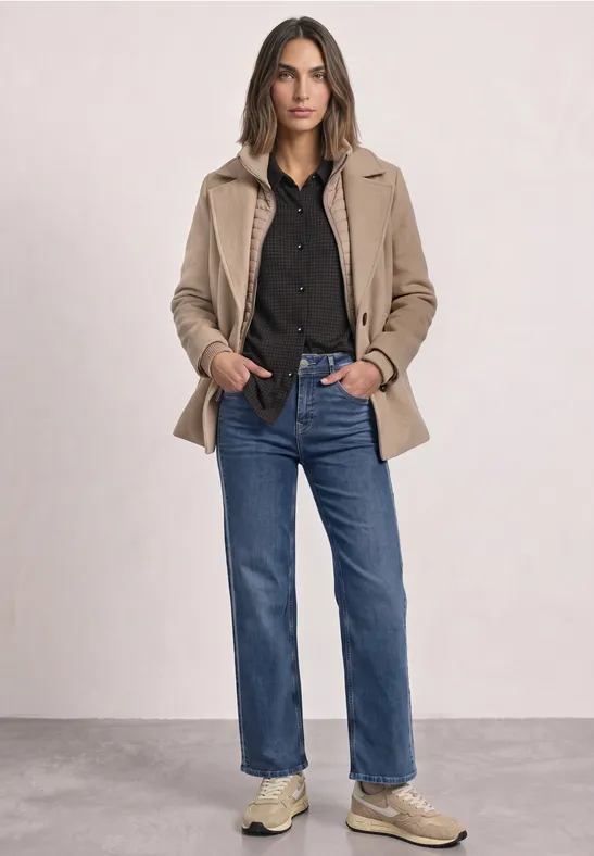 Vrouw met zwart overhemd, beige jas, blauwe jeans en sneakers voor een neutrale achtergrond.
