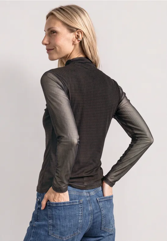 Eine Frau in schwarzem Langarmshirt und Jeans von der Seite, Hände in den Gesäßtaschen.