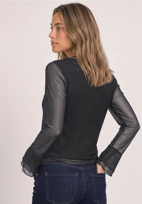 Eine Frau steht seitlich, trägt einen schwarzen Pullover mit transparenten Ärmeln und blaue Jeans.