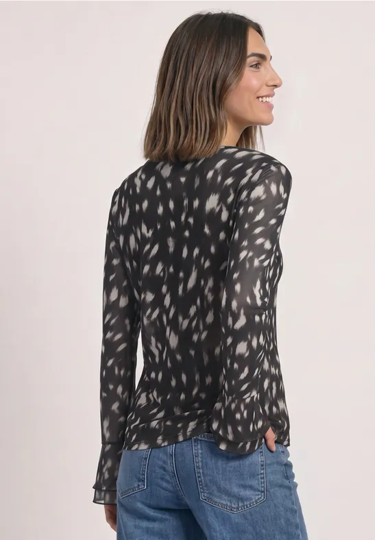 Frau in schwarzer, gemusterter Bluse und Jeans, seitlich fotografiert.