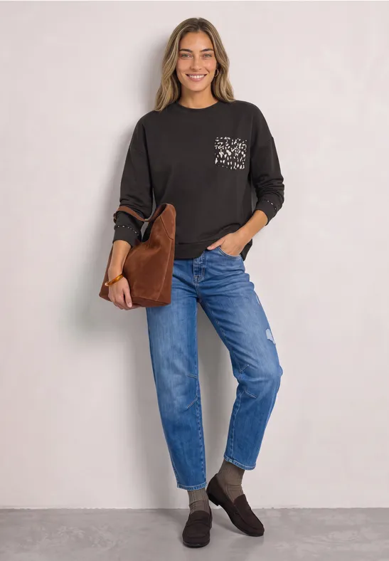 Eine lächelnde Frau trägt ein schwarzes Langarmshirt, blaue Jeans, braune Stiefeletten und eine braune Tasche.