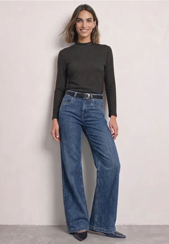 Frau in schwarzem Langarmshirt und weitem Jeans, steht lässig vor hellem Hintergrund.