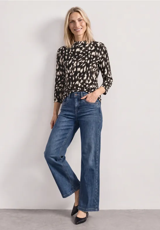 Frau mit schwarzer, gemusterter Bluse, weiter blauer Jeans und schwarzen Pumps vor hellem Hintergrund.