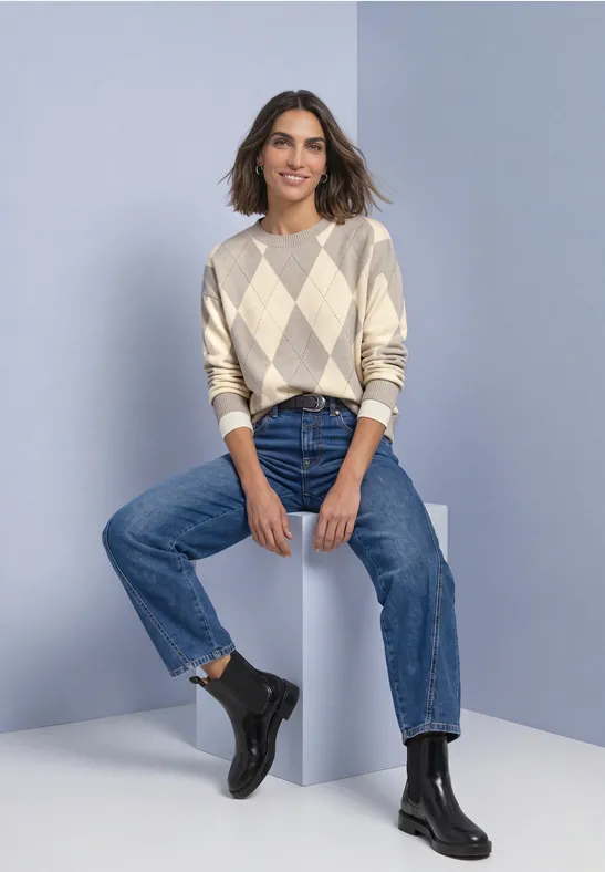 Femme en pull à losanges, jean et bottines est assise détendue sur un cube devant un mur pastel.