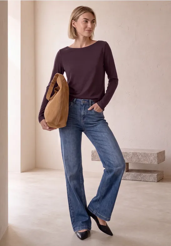 Vrouw in paars langarmshirt, rechte blauwe jeans en zwarte ballerina’s, draagt een bruine tas.