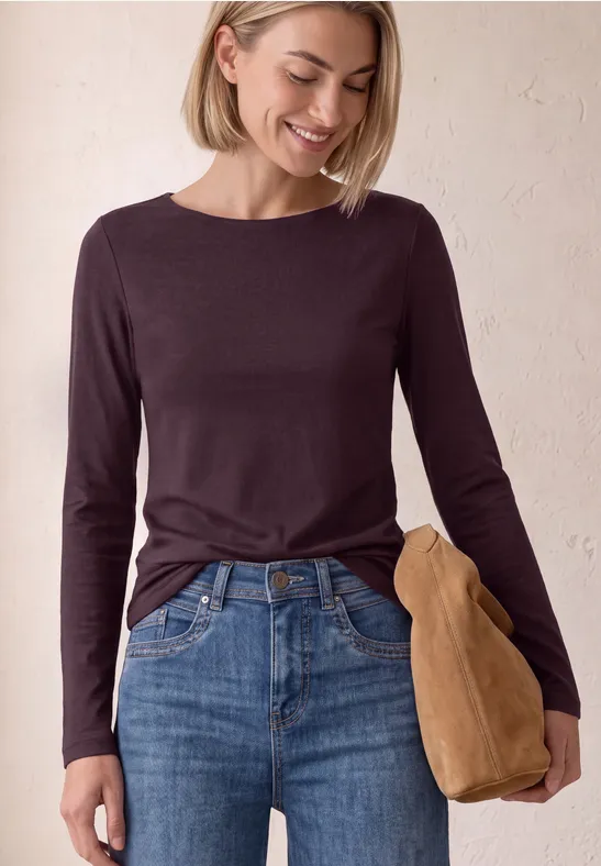 Vrouw in donker lang shirt en spijkerbroek, glimlachend en een bruine tas vasthoudend.