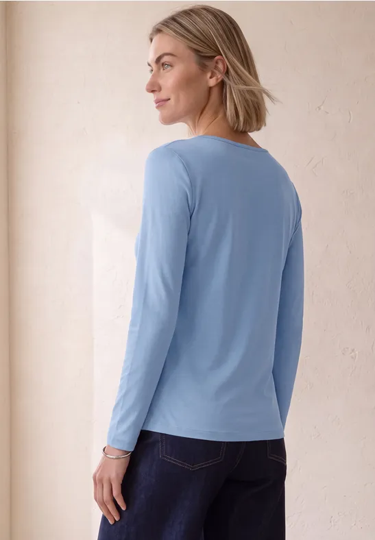 Frau mit schulterlangem blonden Haar trägt ein hellblaues Langarmshirt und blickt seitlich über die Schulter.