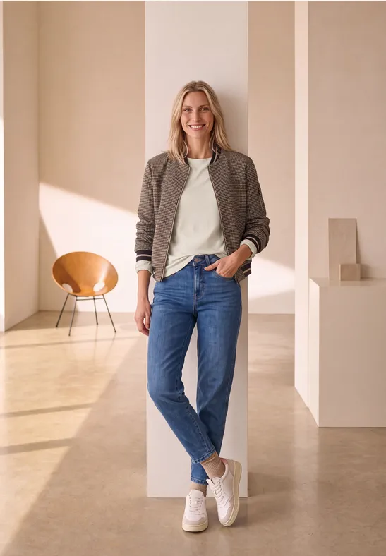 Een lachende vrouw in jeans en sneakers leunt tegen een zuil in een lichte, moderne ruimte.