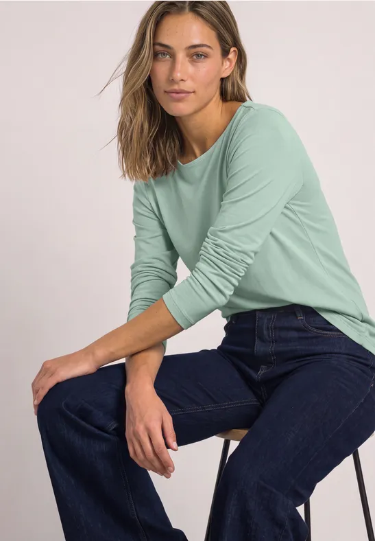 Frau in mintgrünem Langarmshirt und blauen Jeans sitzt lässig auf einem Hocker.