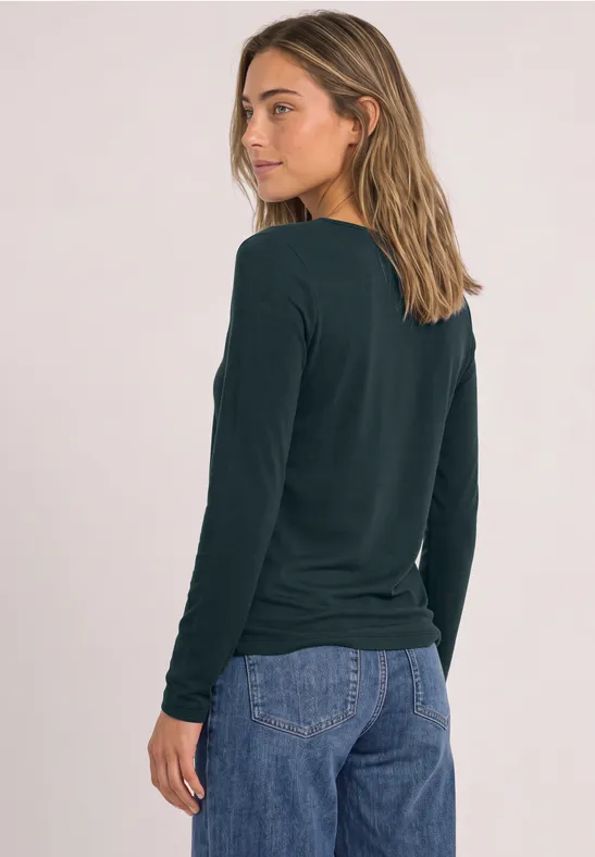 Junge Frau im Profil mit dunkelgrünem Langarmshirt und blauen Jeans vor hellem Hintergrund.