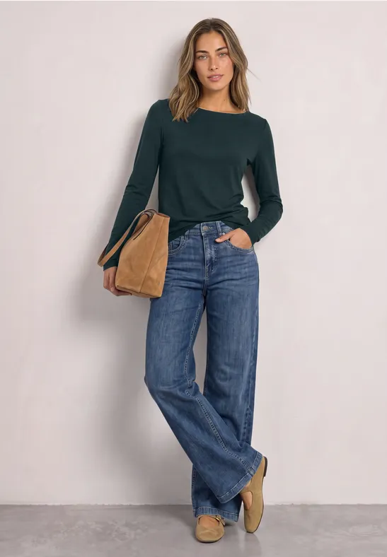 Frau in dunkelgrünem Pullover, weiten Jeans und beiger Tasche vor neutraler Wand.