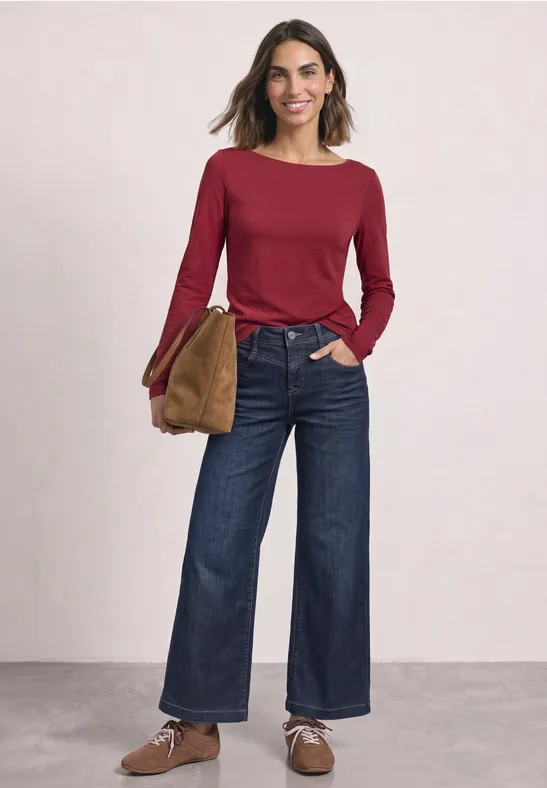 Vrouw in rood shirt met lange mouwen, wijd donkerblauw spijkerbroek, bruine schoenen en tas tegen lichte achtergrond.
