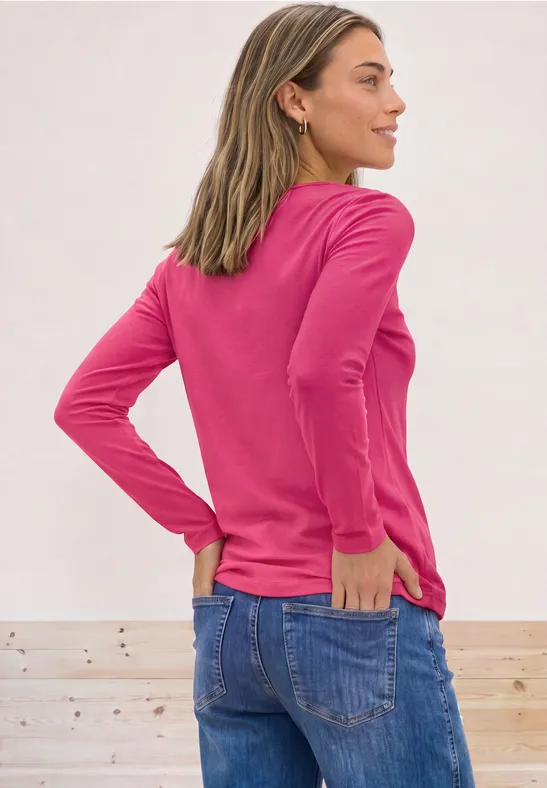 Vrouw van achteren in roze longsleeve en spijkerbroek, handen in de zakken, lichte achtergrond.