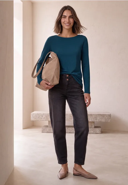 Frau in blaugrünem Langarmshirt, schwarzer Jeans und beigen Ballerinas mit Tasche vor schlichtem Interieur.
