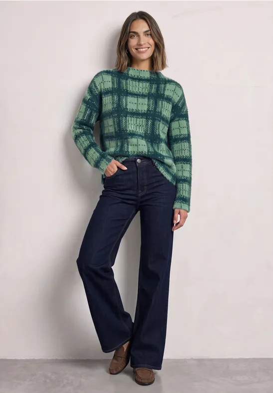 Een vrouw draagt een groene geruite trui en wijde jeans tegen een lichte achtergrond.