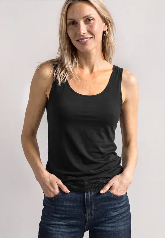 Lächelnde Frau trägt schwarzes Top und Jeans, steht vor hellem Hintergrund mit Händen in den Taschen.