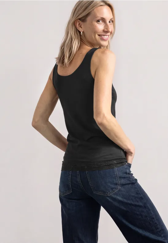 Frau in schwarzem Top und blauen Jeans, lächelnd, leicht zur Kamera gedreht, Hände in den Gesäßtaschen.