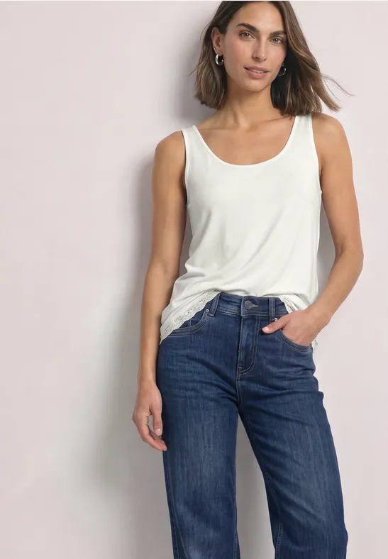 Eine Frau steht lässig mit einer Hand in der Jeanstasche, trägt weißes Tanktop und blaue Jeans.